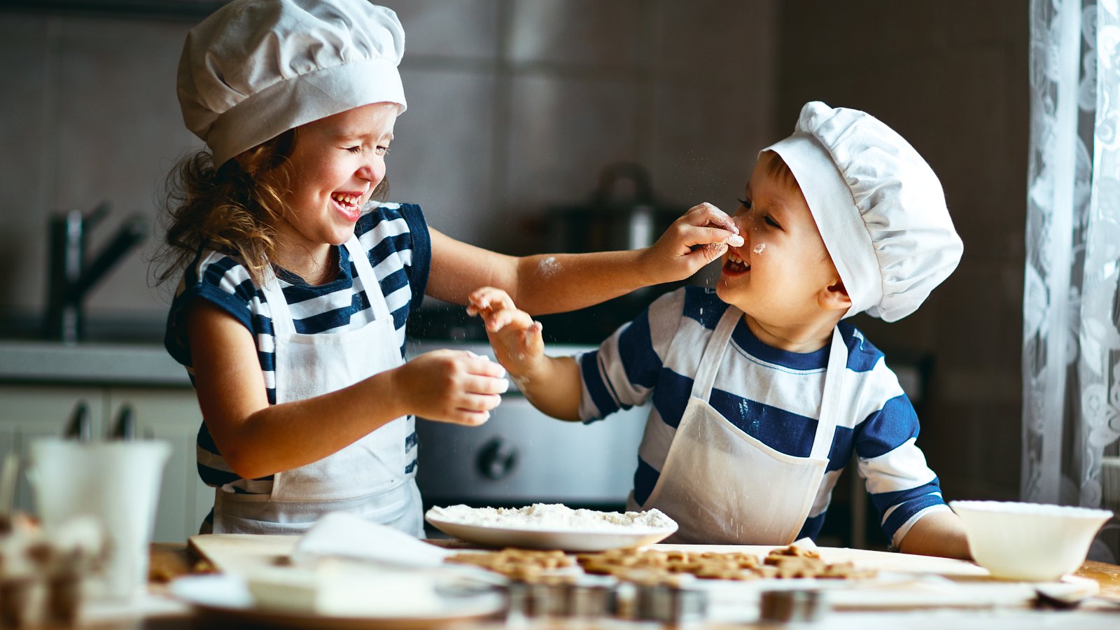 2 lachende kleine kinderen met koksmutsen op, smeren meel op elkaars neusje