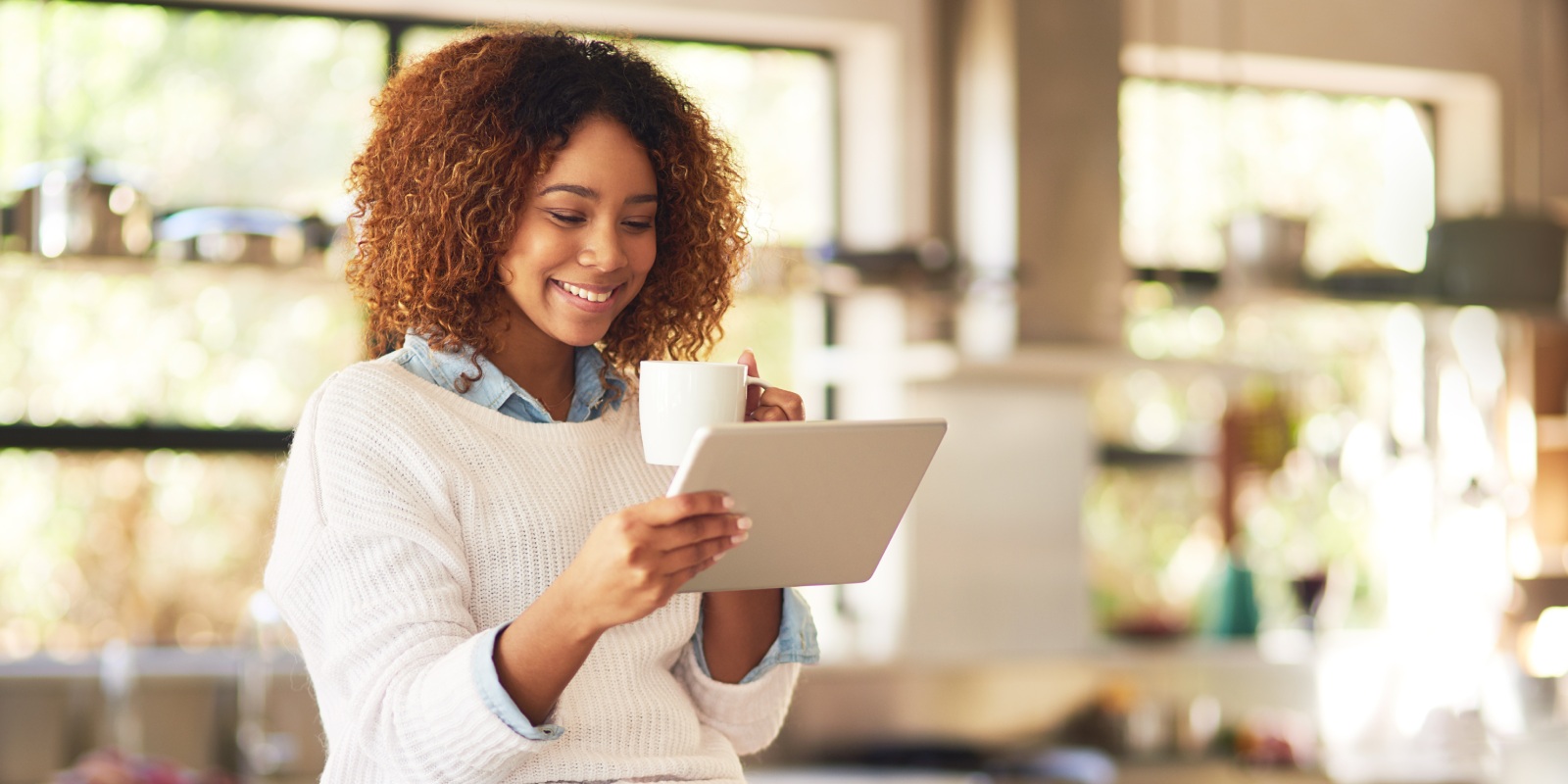 Blije vrouw met bruin haar en witte trui drinkt koffie en bekijkt op haar tablet de Jaarcijfers zorg 2023 