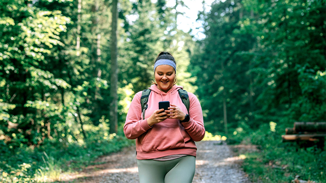 Een vrouw in sportieve kleding wandelt vrolijk door het bos met een rugzak op. Met een brede lach kijkt ze op haar telefoon. Ze loopt recht op je af en lijkt klaar voor een actieve tocht.